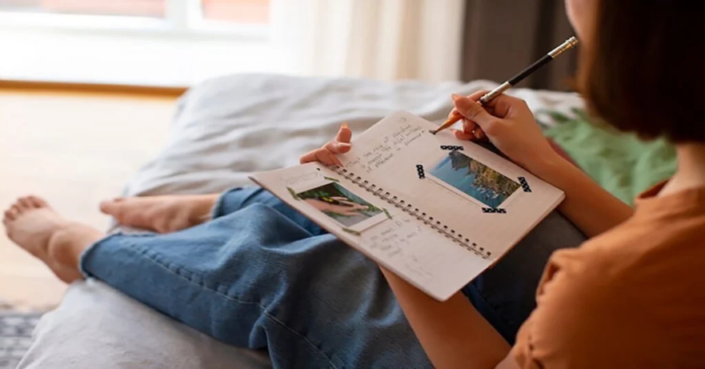 A person sitting on a bed writing in a simple journaling notebook with attached travel photos, capturing memories and reflections.
