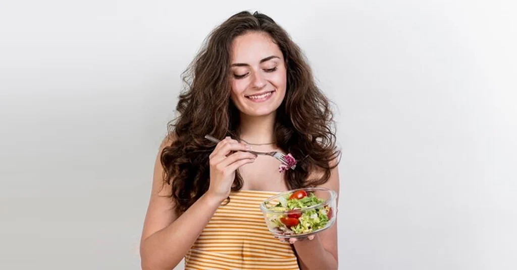 A woman smiling while enjoying a healthy salad, promoting physical self-care routines.
