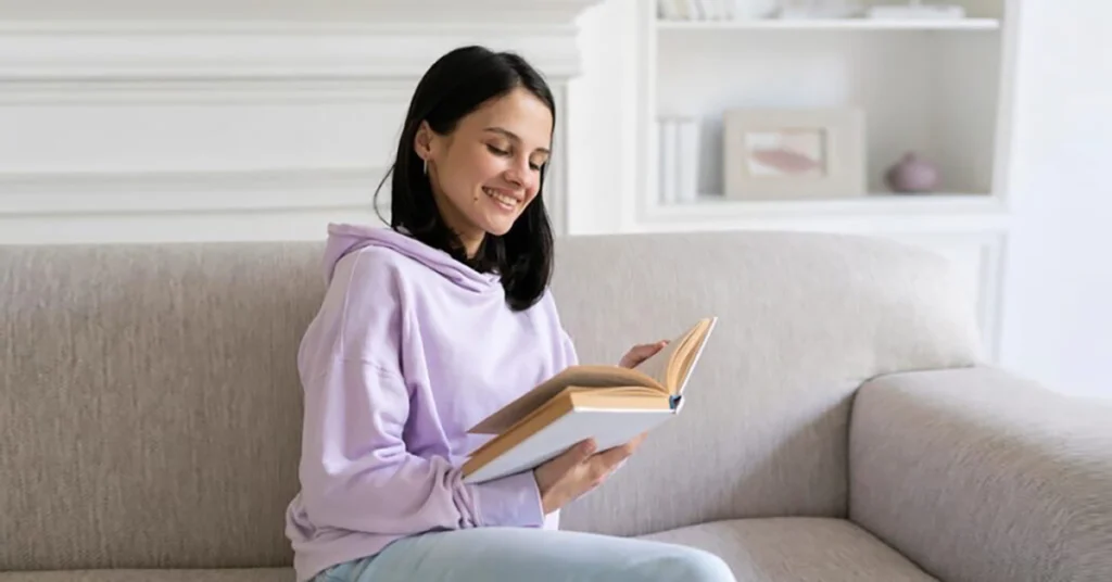  A woman is relaxing on a couch, smiling as she reads a book as part of her self-care routine. 
