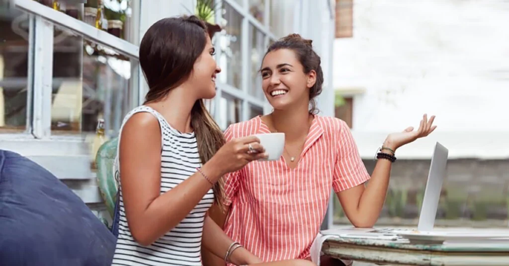 Two women are sitting outdoors, laughing and talking while one holds a coffee cup.