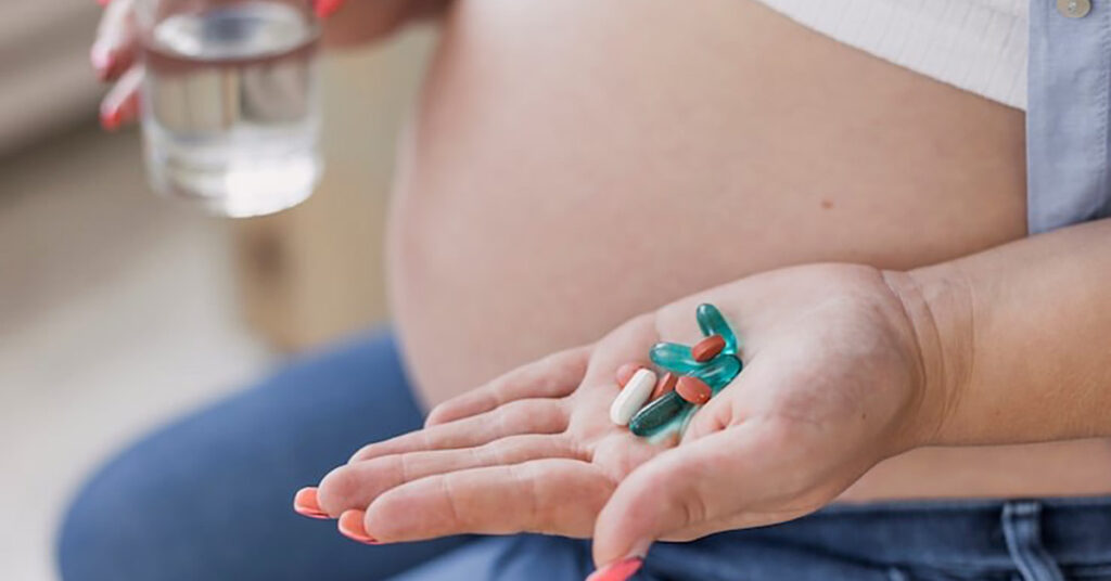 Pregnant woman holding multiple pills including Tylenol in hand while holding glass of water, focusing on safe medicine use.